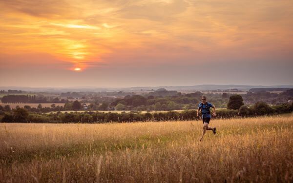 A person running through a meadow at sunset with a view of trees, hills and fields in the background