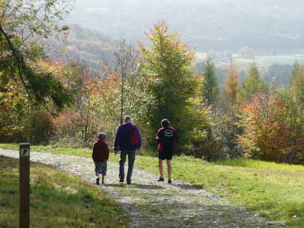 Two adults and a child walk along a pathway with Autumn trees and a valley in the background