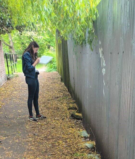 A girl with a clipboard stands on a path by a big grey fence