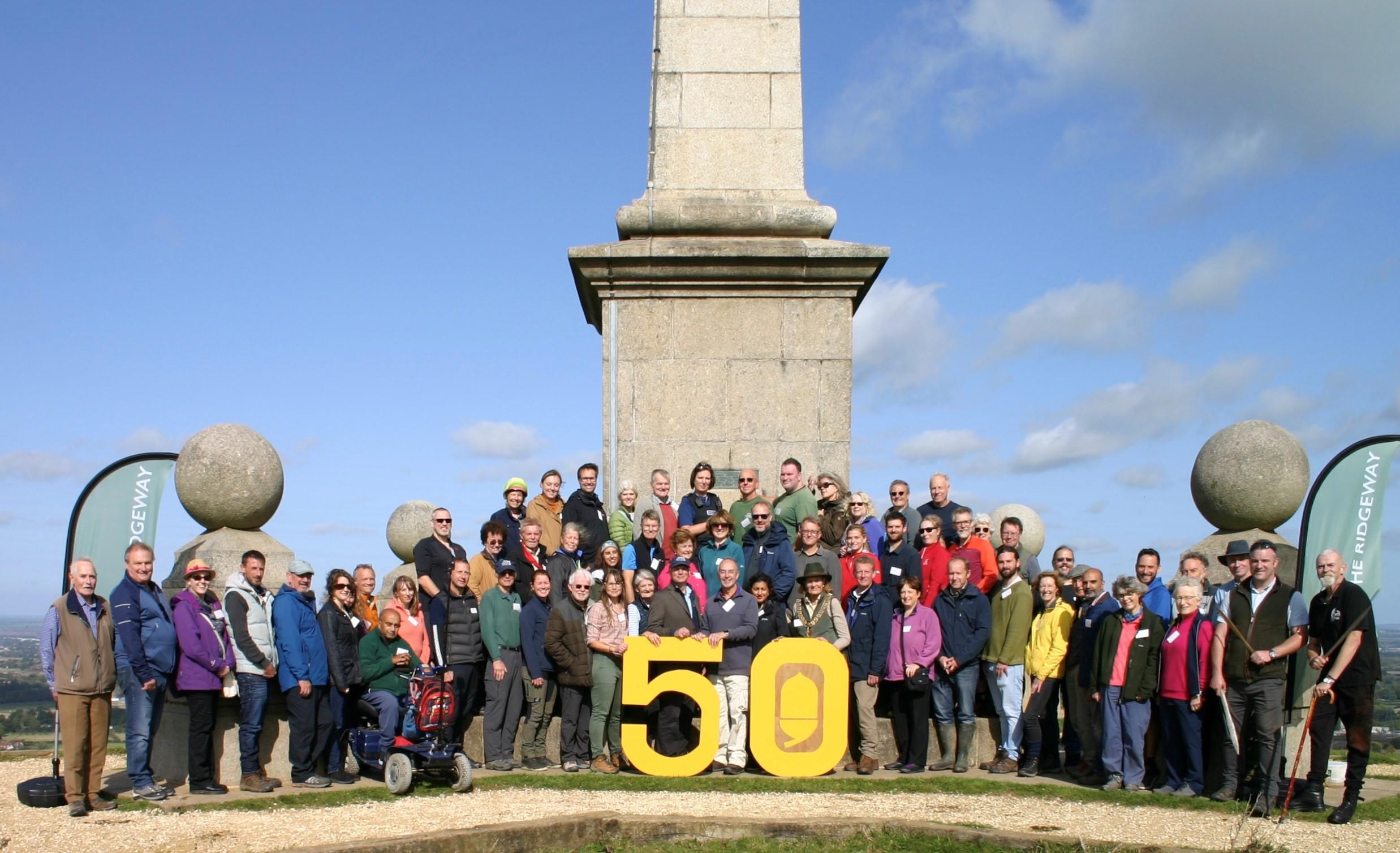 A group of about 70 people stand in front of the monument at the top of Coombe Hill with a big yellow 50 sign.