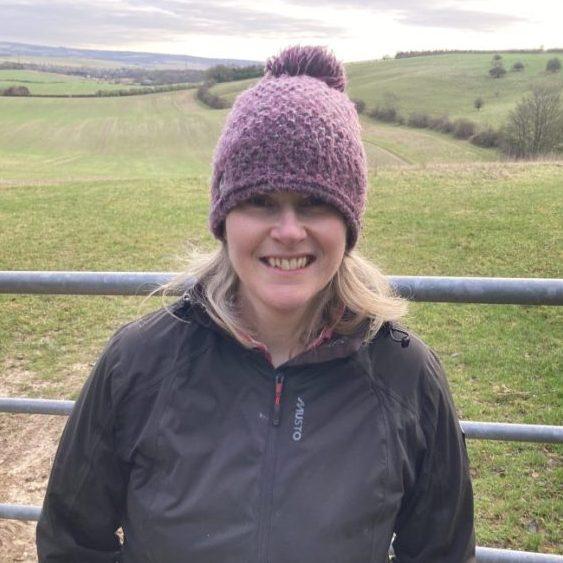 Photo of a woman with blonde hair, wearing a purple bobble hat, standing in a field.