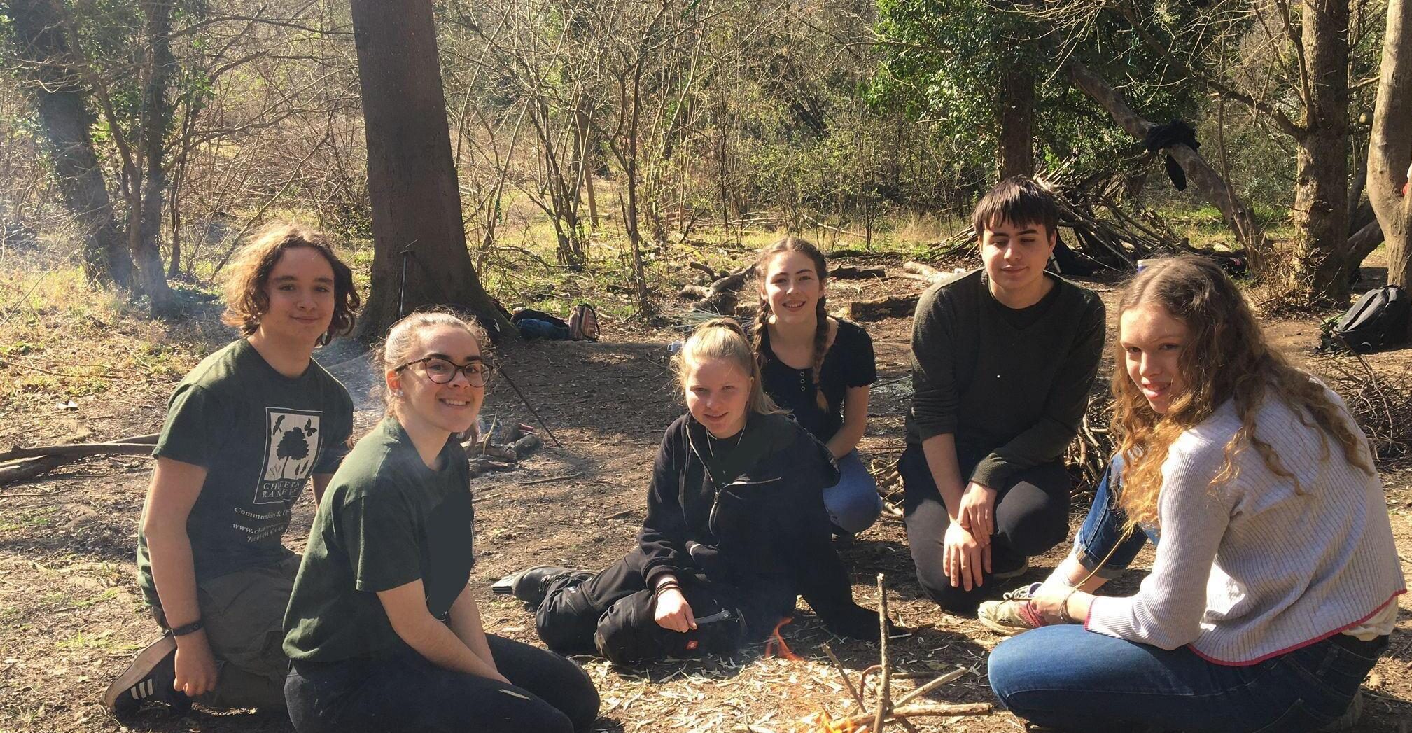 A group of teens sit around a campfire in the woods.
