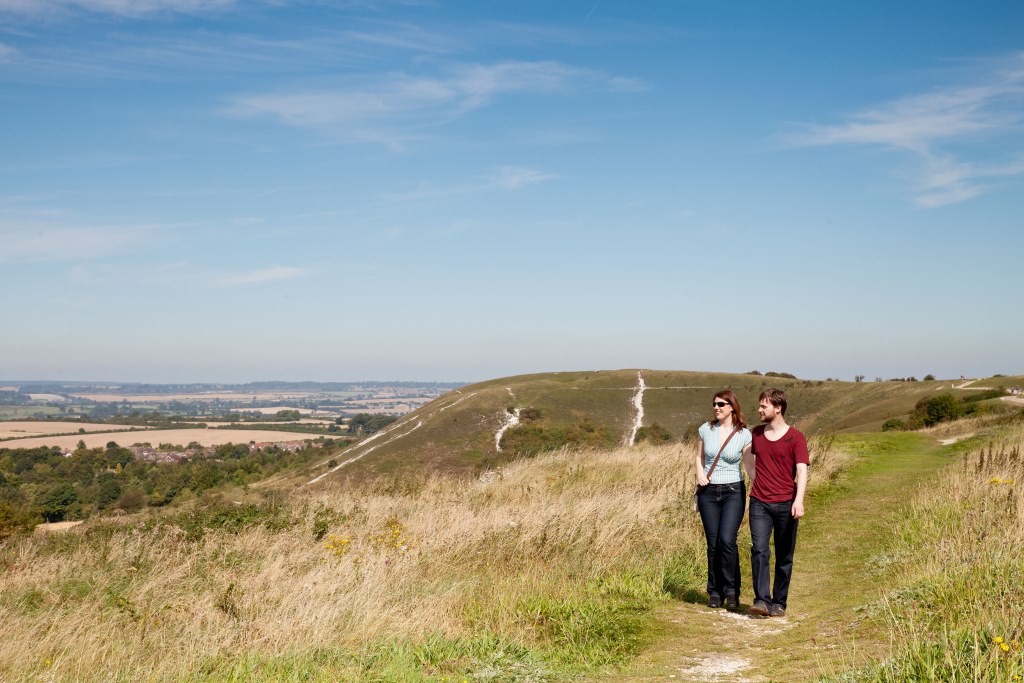 A man and woman walk along a grass path on a hill with views of fields behind them.