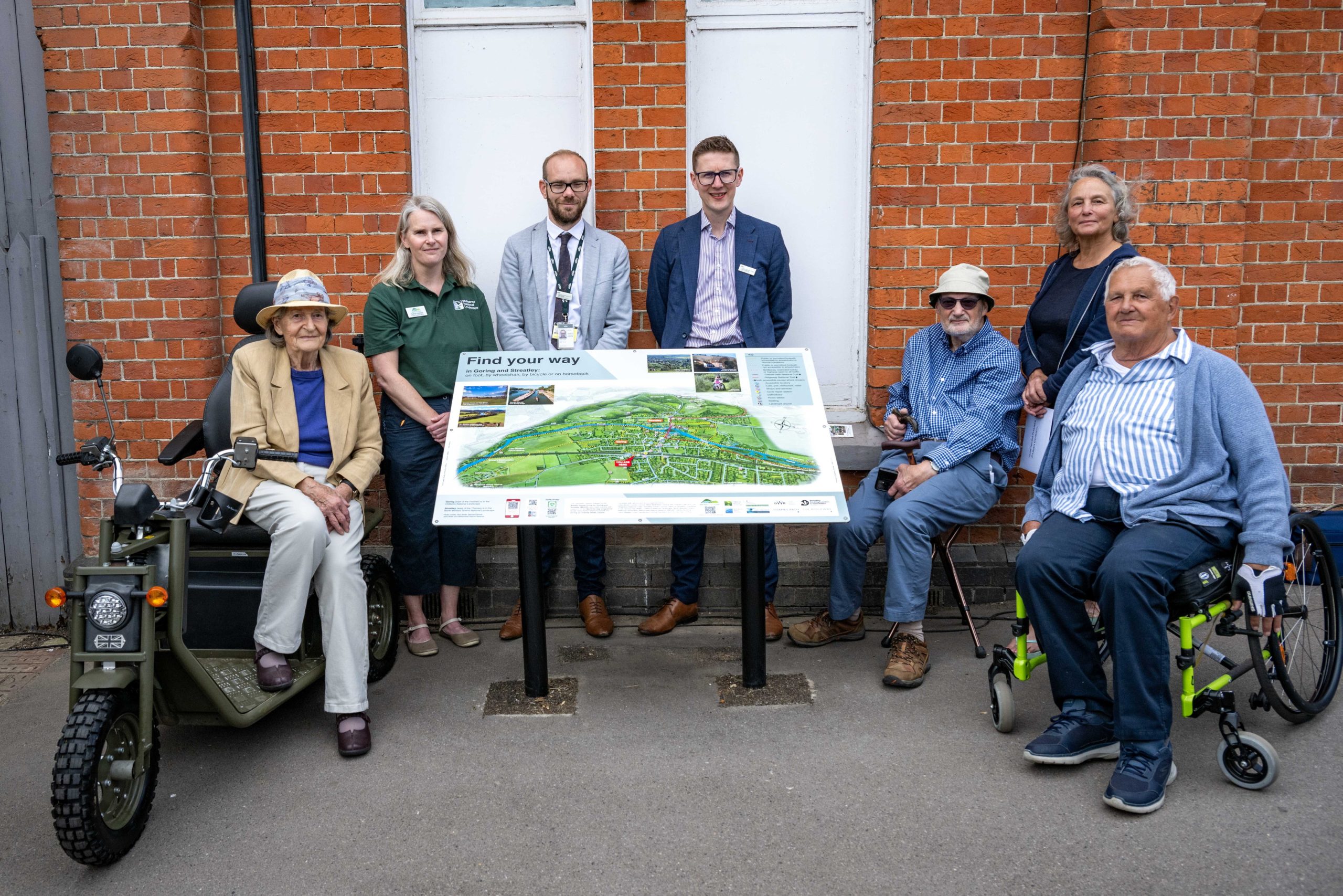A group of people around an information board. Four people stand behind the board and three people in mobility scooters sit next to it.