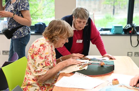 A woman sits in front of a cushion with lace-making bobbins while another woman watches.