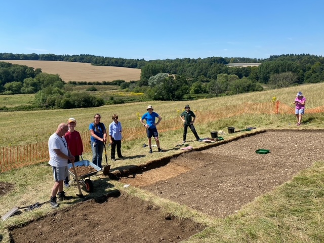 A group of people stand on the edge of a rectangle dug into the earth.