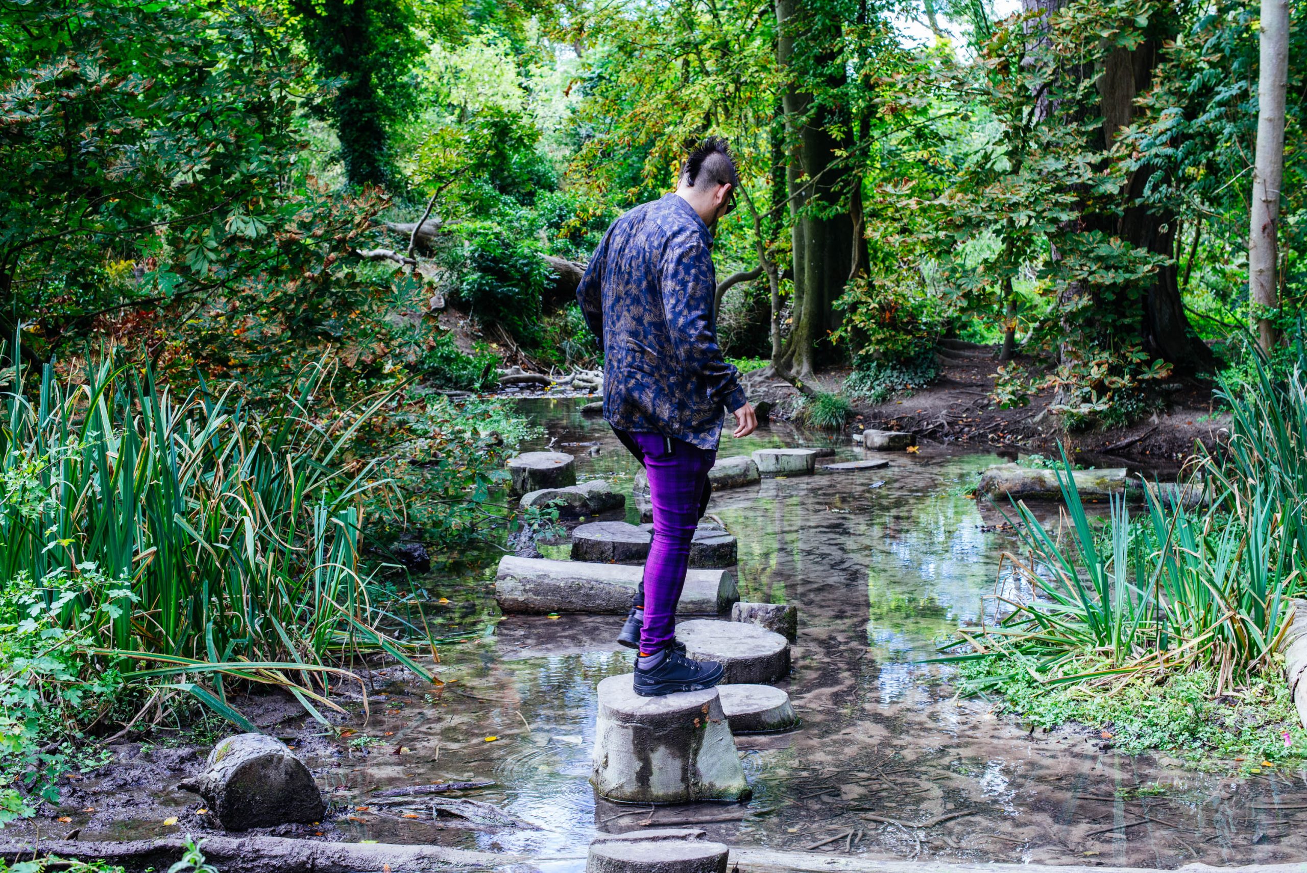 A male figure walking along stepping stones in a stream surrounded by lush green vegetation and trees. The male is facing away from the camera.