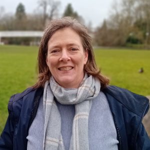 A woman standing in a field wearing a dark blue jacket and light blue scarf.