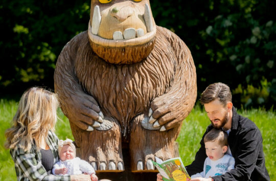 woman with baby sits by gruffalo statue while man reads to toddler from gruffalo book